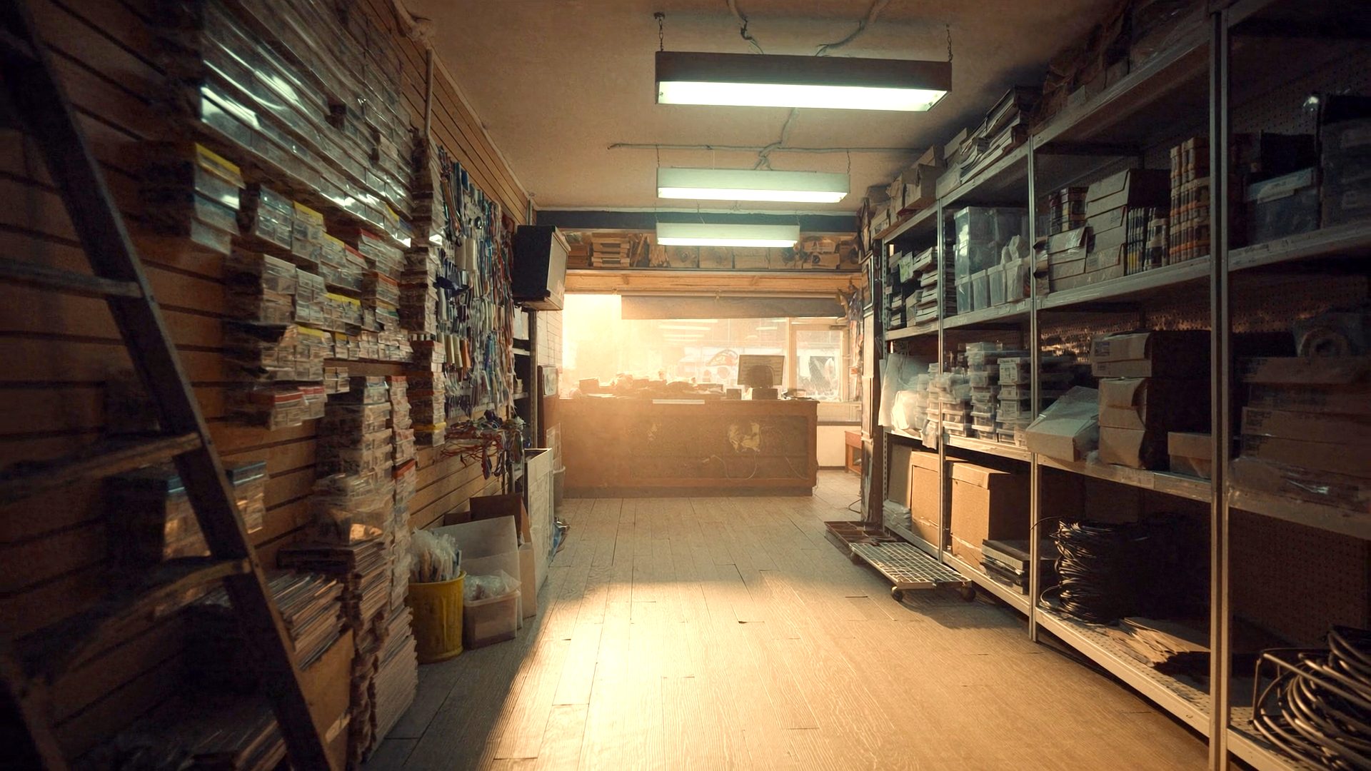 An aisle in the store looking toward the service counter, work lights on and late sun through the front window.