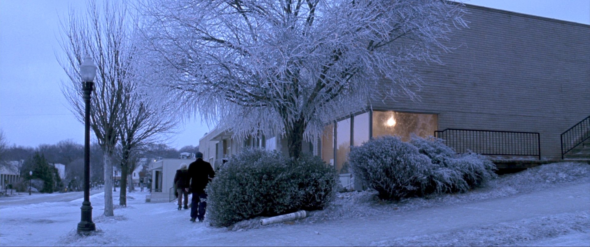 Main Street glazed in ice at first light, the trees bent under it, two people walking the sidewalk, warm light on inside one low brick storefront.